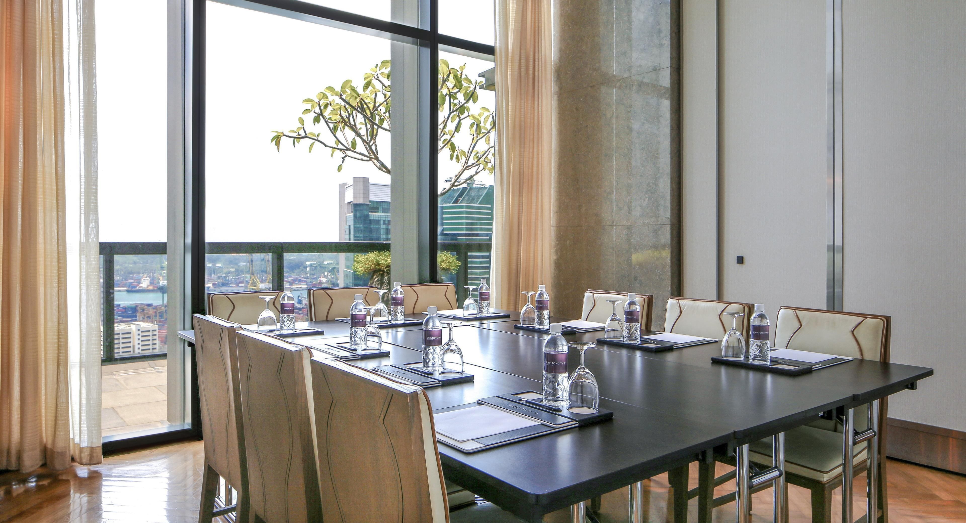 Un salón de conferencias con una larga mesa, sillas, botellas de agua y vistas a un paisaje urbano a través de amplias ventanas.