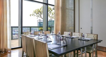 A conference room with a long table, chairs, water bottles, and a view of a cityscape through large windows.
