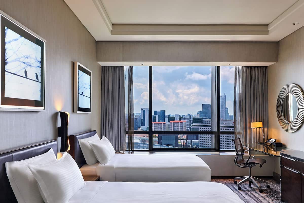 A hotel room with two beds, a desk, a chair, and a large window offering a city view.