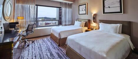A hotel room with two beds, a desk with a chair, a coffee maker, and a view of buildings through the window.