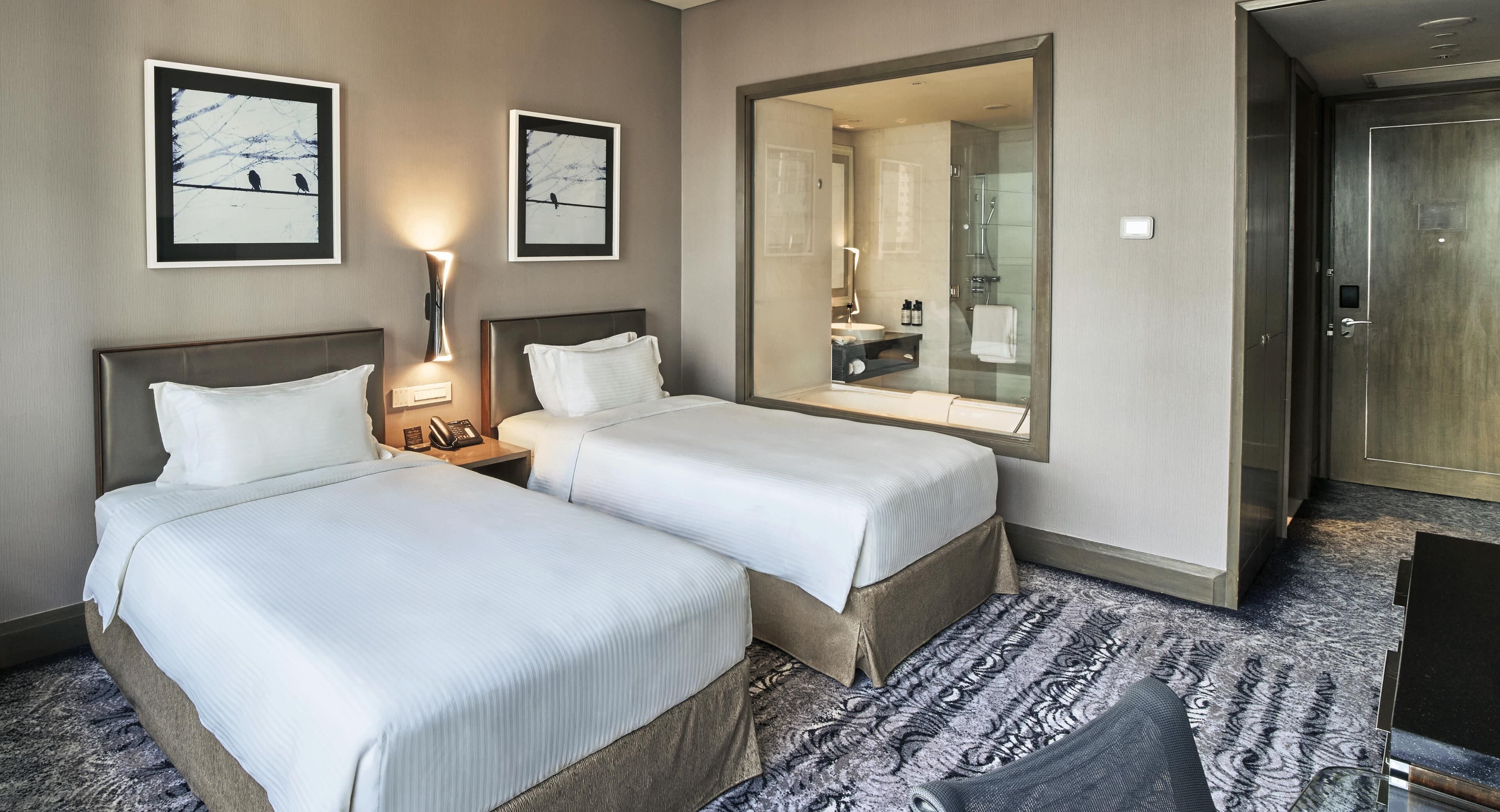 Hotellihuone, jossa on kaksi sänkyä, yöpöytä lampun kanssa, suuri peili ja suihkutila.