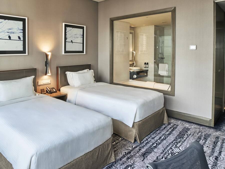 Hotellihuone, jossa on kaksi sänkyä, yöpöytä lampun kanssa, suuri peili ja suihkutila.