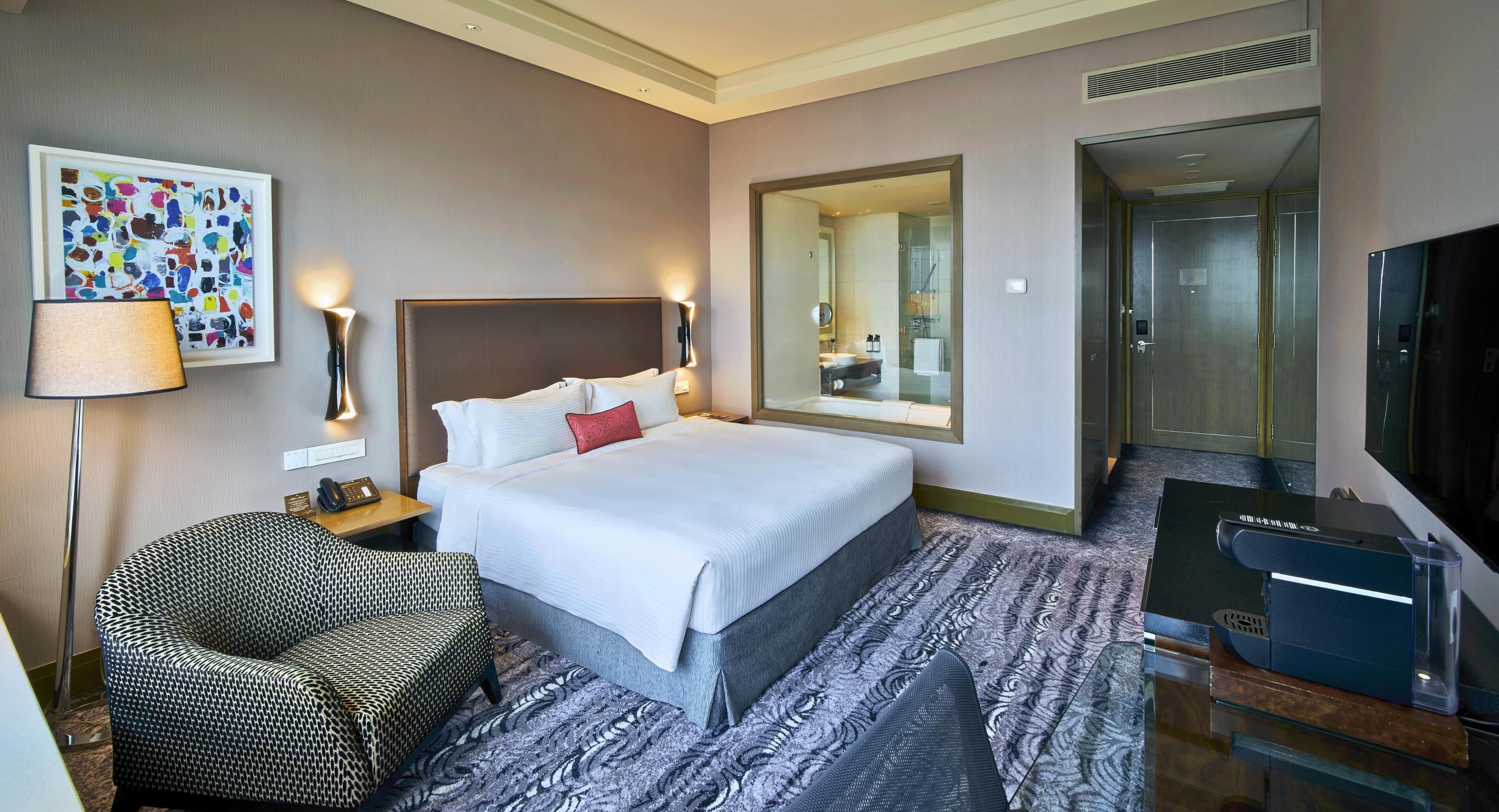 A hotel room with a bed, a chair, a desk with a computer, a TV, and a bathroom visible through a glass door.