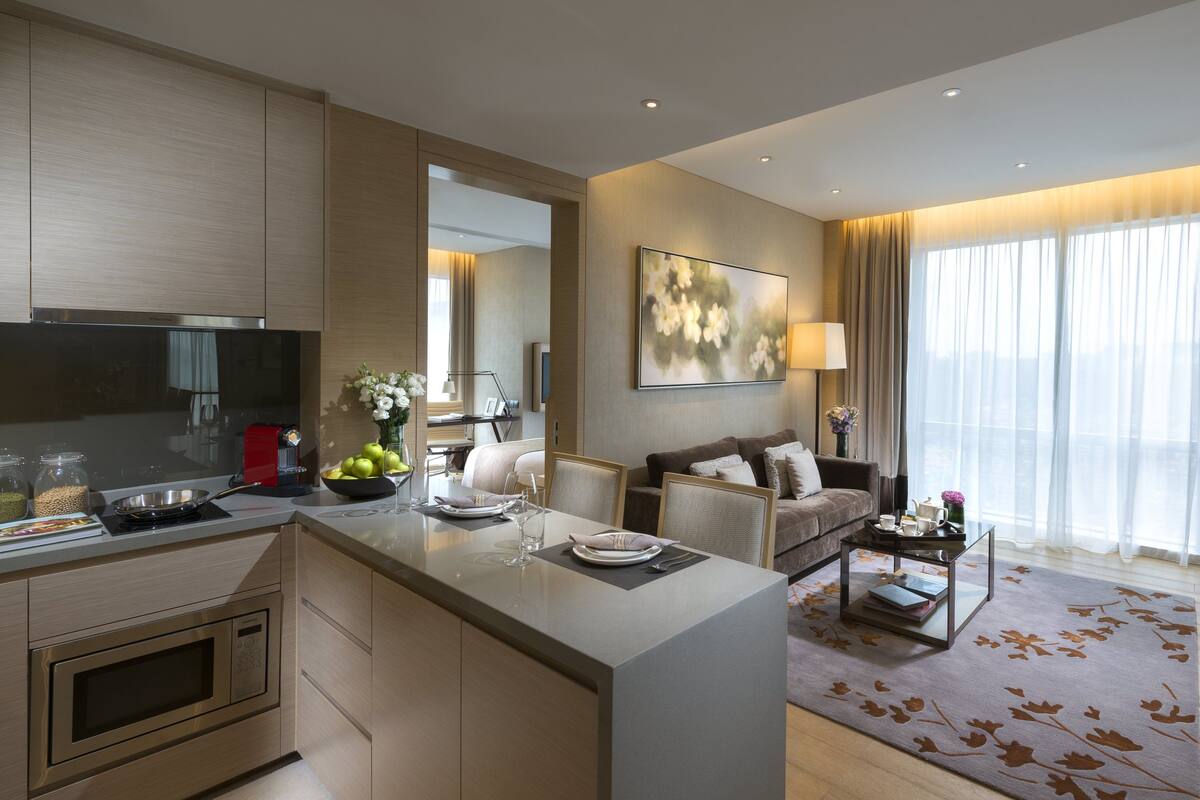 Ett modernt hotellrum med ett litet kök, ett matbord för två personer, en soffa, en TV och ett stort fönster med gardiner.