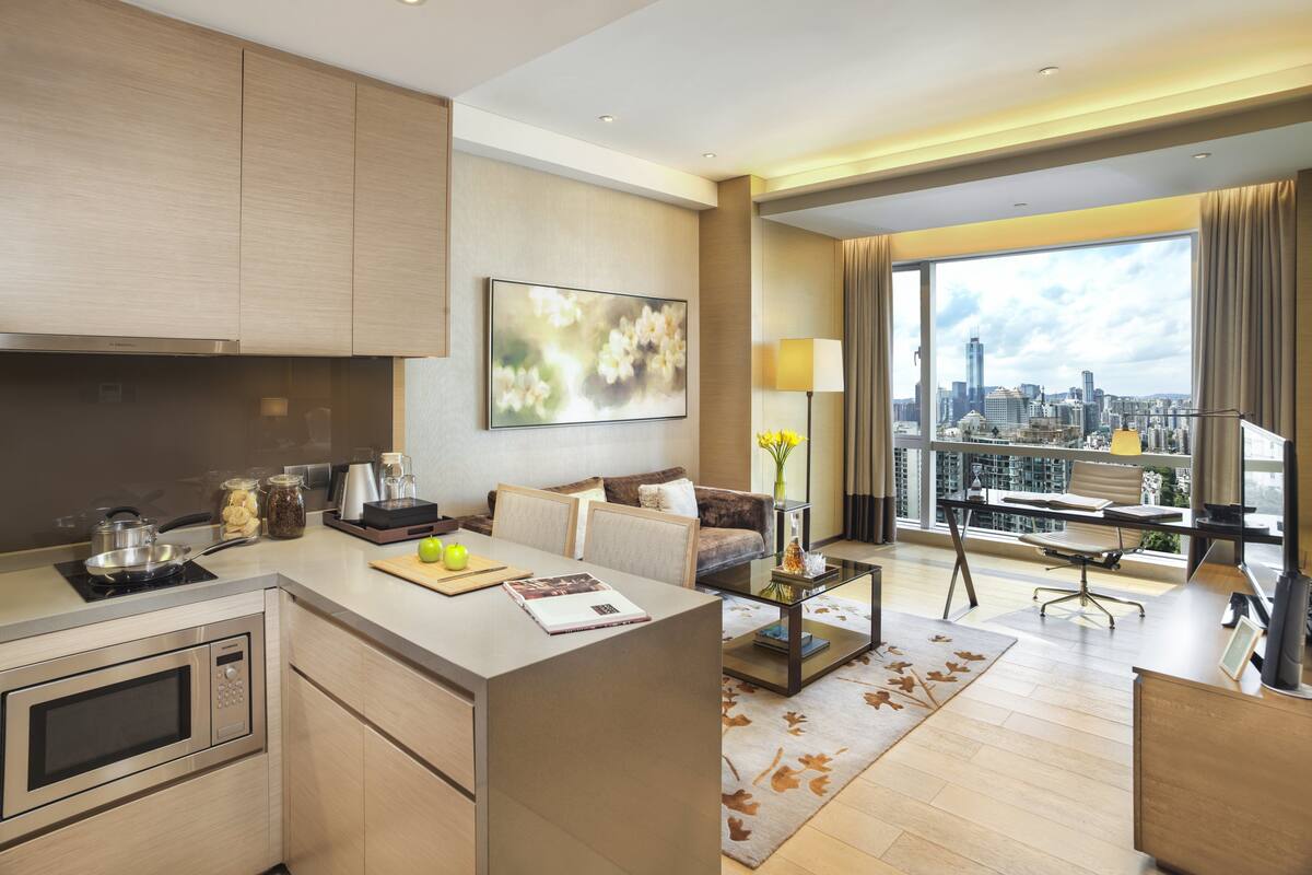 Ett modernt hotellrum med ett kök, ett vardagsrum med soffa och soffbord, en matplats med skrivbord och stol samt ett stort fönster med utsikt över staden.