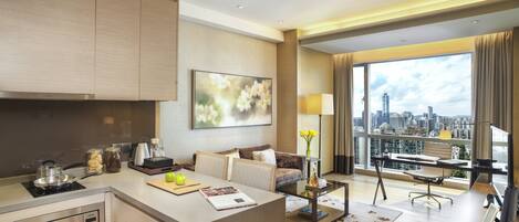 A modern hotel room with a kitchen, a living area with a sofa and coffee table, a dining area with a desk and chair, and a large window with a city view.