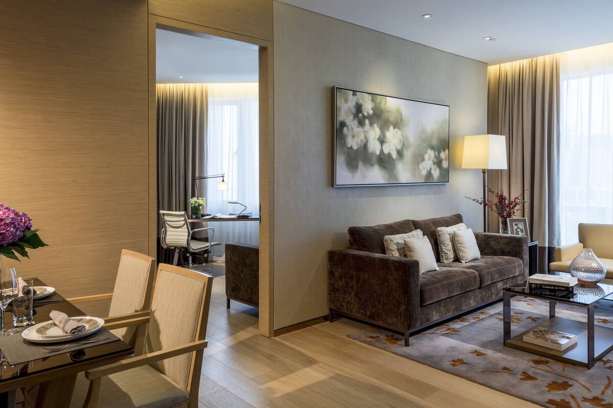Ett modernt hotellrum med en brun soffa, ett matbord för två personer, en stor tavla på väggen och utsikt över staden genom stora fönster.
