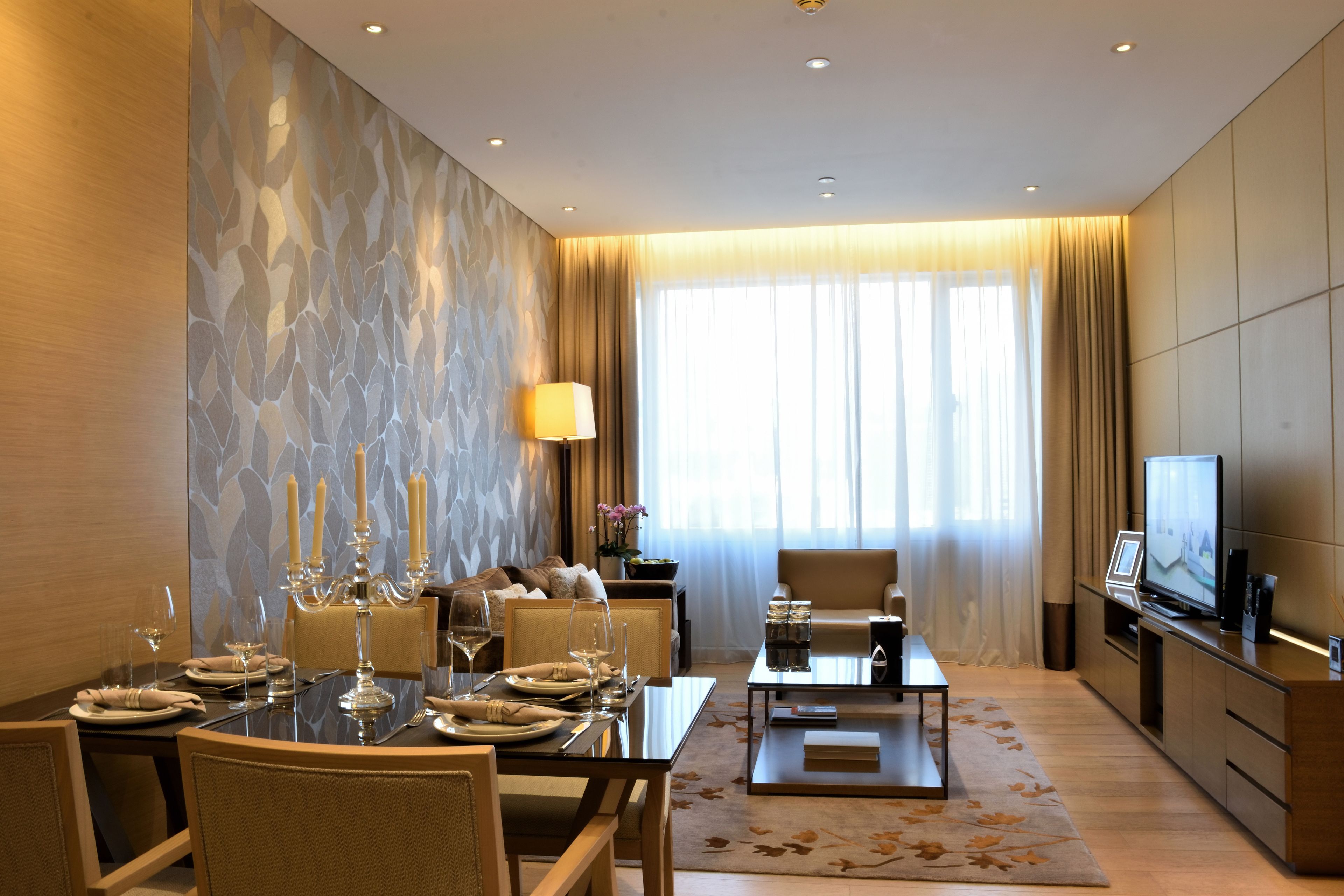 A modern hotel room with a dining area, a sofa, a television, and a large window with curtains.