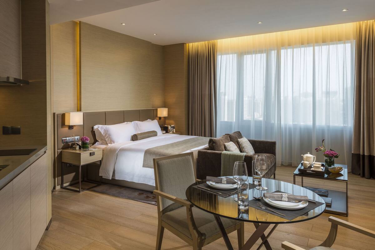 Ett hotellrum med en säng, sängbord, ett glasbord och utsikt över omgivningarna genom stora fönster.