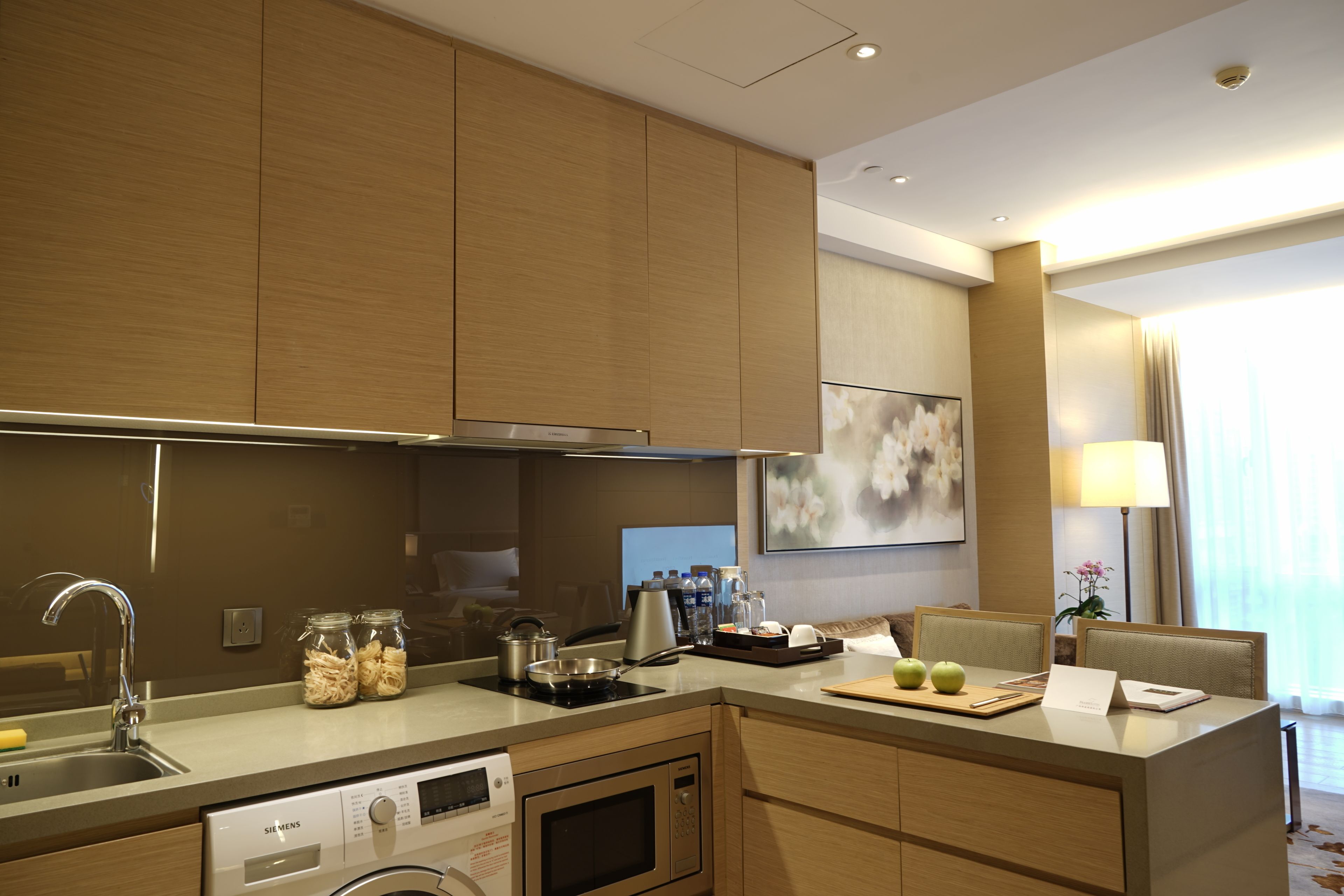 A modern kitchen with stainless steel appliances, a built-in dishwasher, and a microwave. There are two jars of pasta on the counter, a kettle, and a tray with a teapot. The kitchen has wooden cabinets and a large window with curtains.