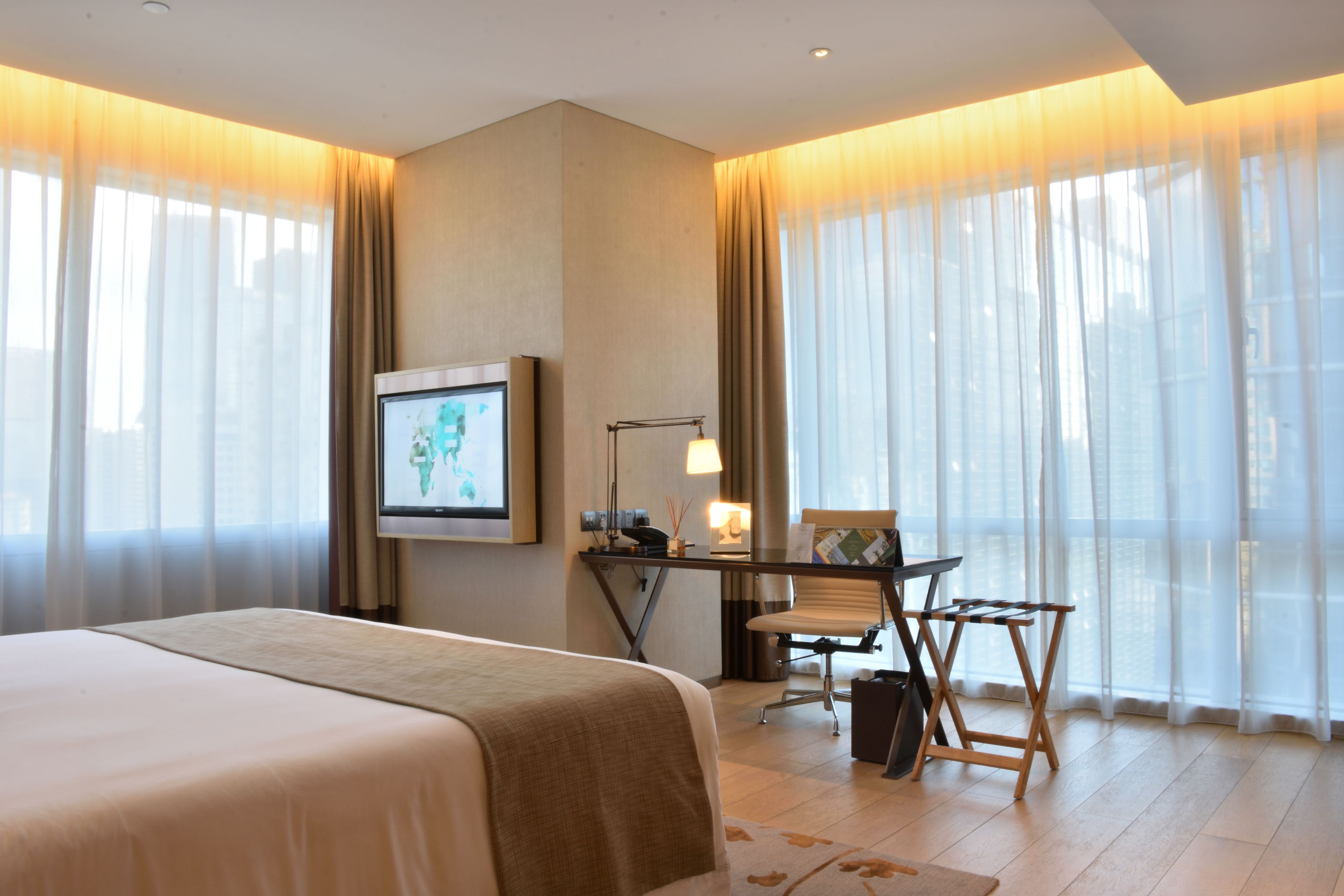 Habitación de hotel con una cama grande, un escritorio con silla, televisión y vistas a través de cortinas traslúcidas.