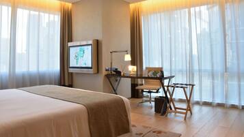 Habitación de hotel con una cama grande, un escritorio con silla, televisión y vistas a través de cortinas traslúcidas.