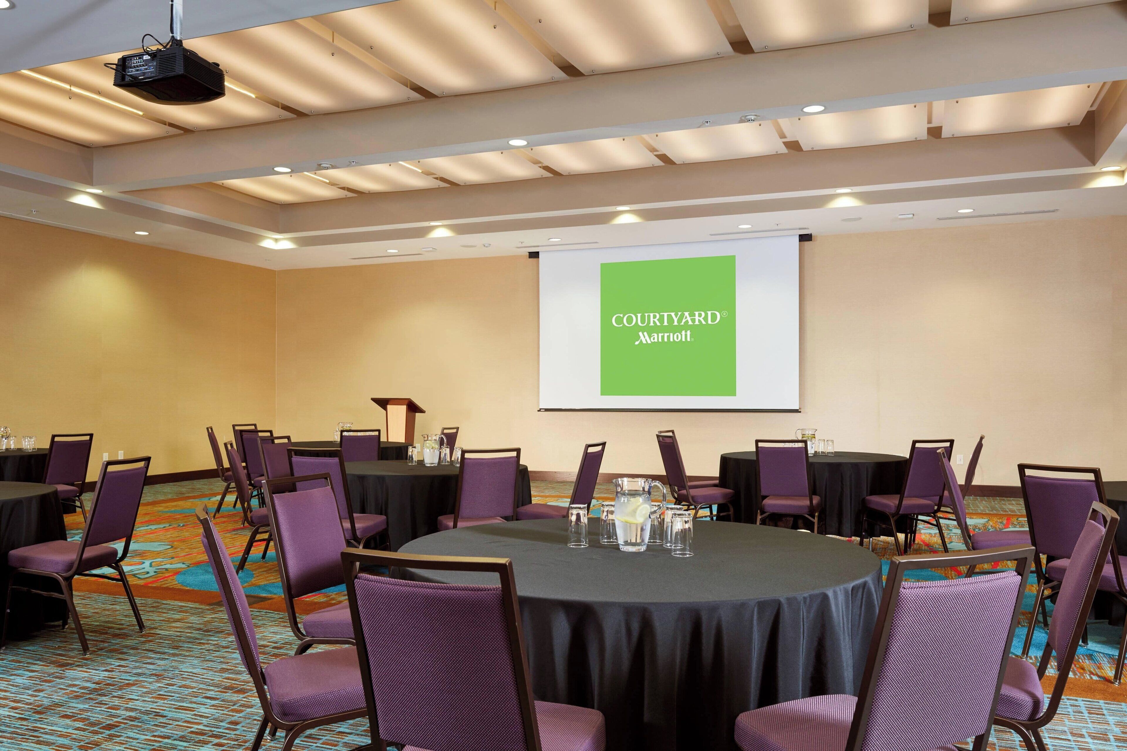 Un salón de conferencias con mesas redondas, sillas moradas y una pantalla grande que muestra "Courtyard Marriott".
