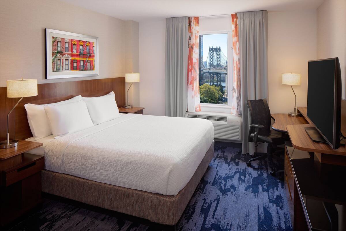 Ein Hotelzimmer mit einem großen Bett, einem Schreibtisch mit Computer, einem Fernseher und einem Fenster mit Vorhängen.