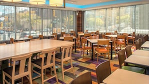 Une salle à manger d’hôtel avec de nombreuses tables et chaises, de grandes fenêtres avec des rideaux et une moquette colorée.