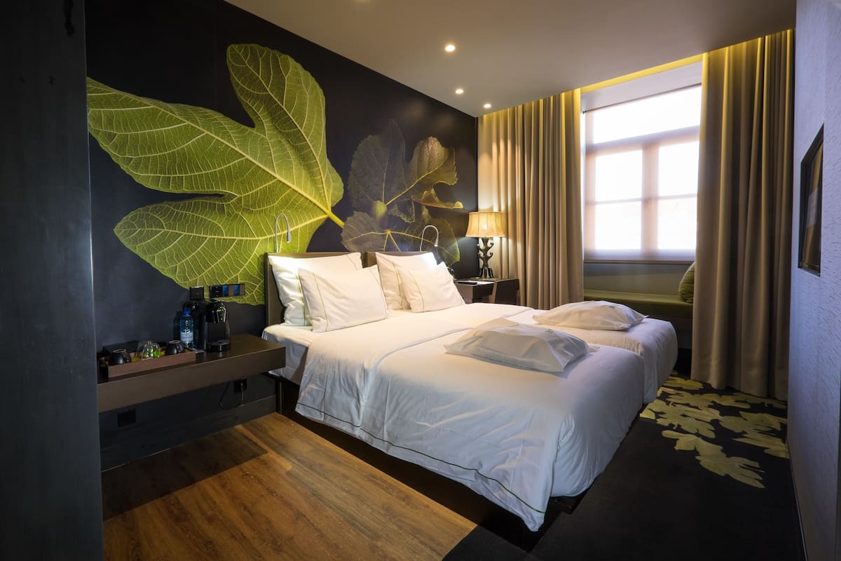 Une chambre d’hôtel avec une grande fresque représentant des feuilles, un lit avec du linge de maison blanc, une table de chevet avec une lampe et une fenêtre avec des rideaux.