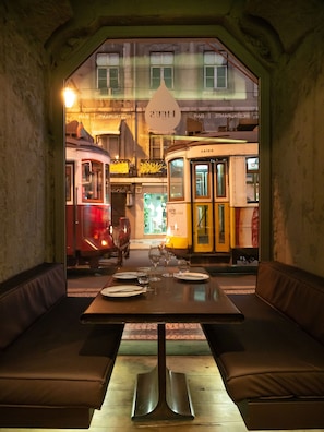 Comedor con una mesa dispuesta para dos, un banco y vistas a tranvías antiguos que circulan.