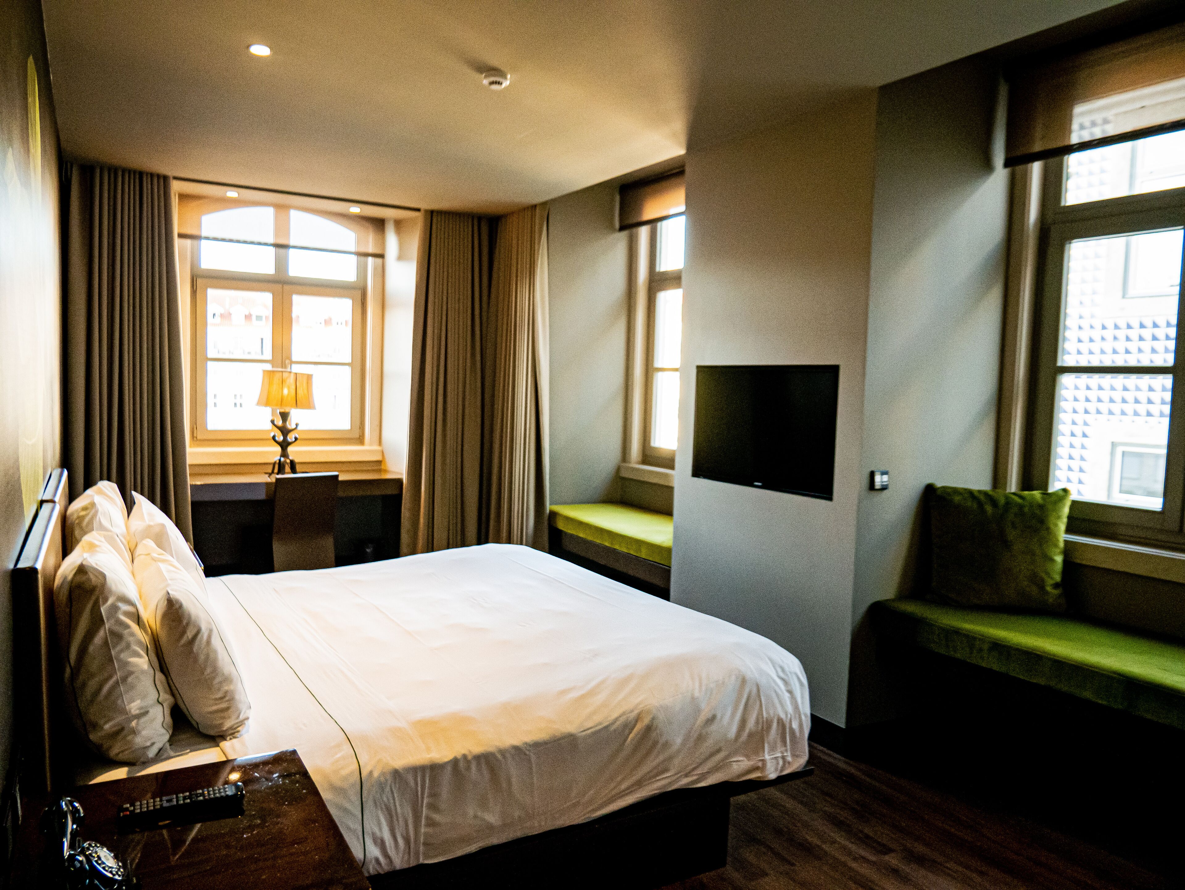 Habitación de hotel con una cama grande, un escritorio con lámpara, un televisor en la pared y una ventana con cortinas.