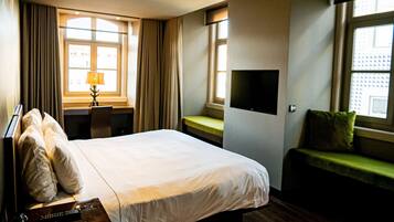 Habitación de hotel con una cama grande, un escritorio con lámpara, un televisor en la pared y una ventana con cortinas.