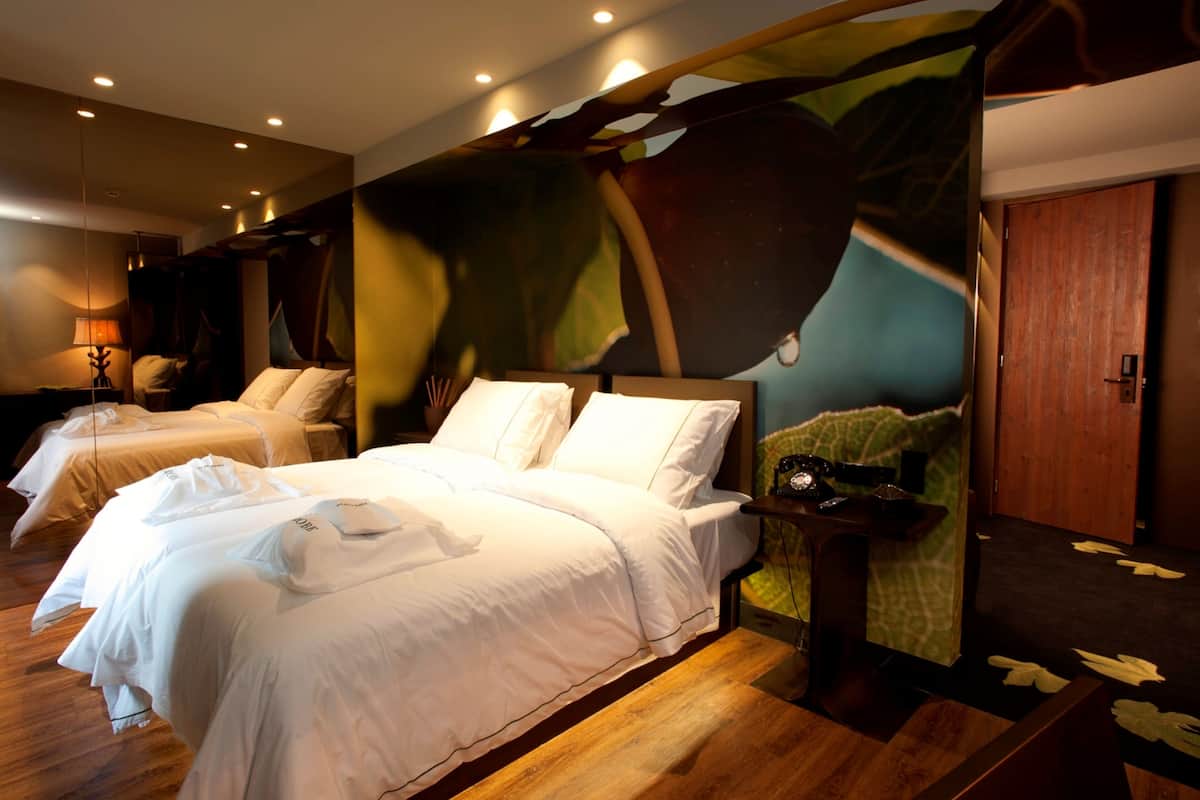 Une chambre d’hôtel avec un grand lit, un plus petit lit et un banc. Il y a un tableau au mur et une porte en bois.