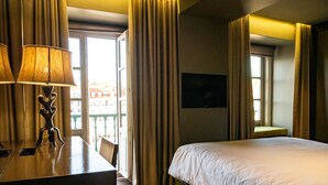 A hotel room with a bed, a desk with a lamp, a chair, a TV on the wall, and a window with curtains. - Figueira by The Beautique Hotels & SPA (Lisbon)