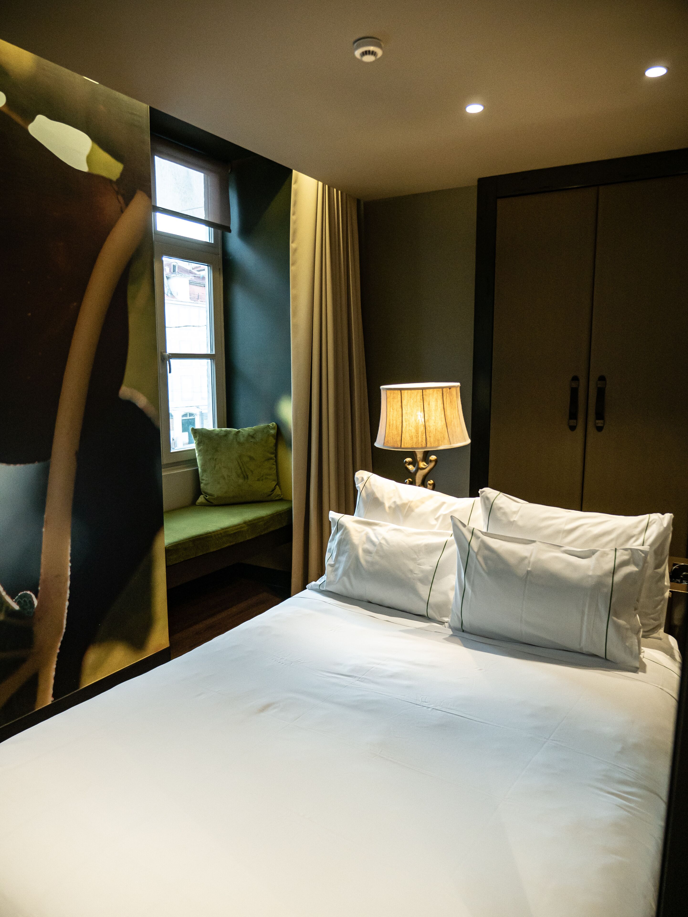 A hotel room with a bed, a lamp, a green chair, and a wardrobe.