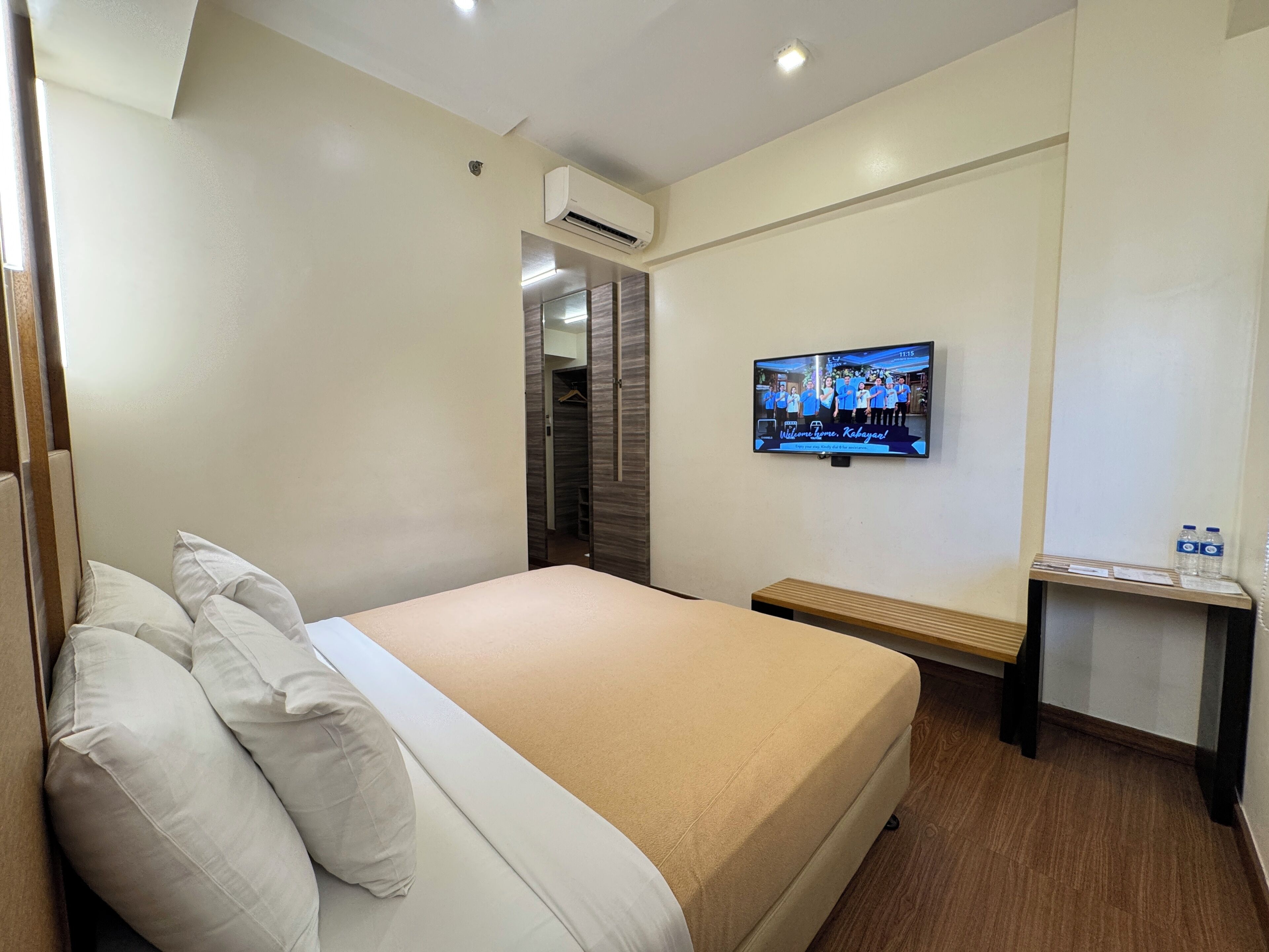A hotel room with a bed, pillows, a TV mounted on the wall, an air conditioning unit, and a small table with water bottles.