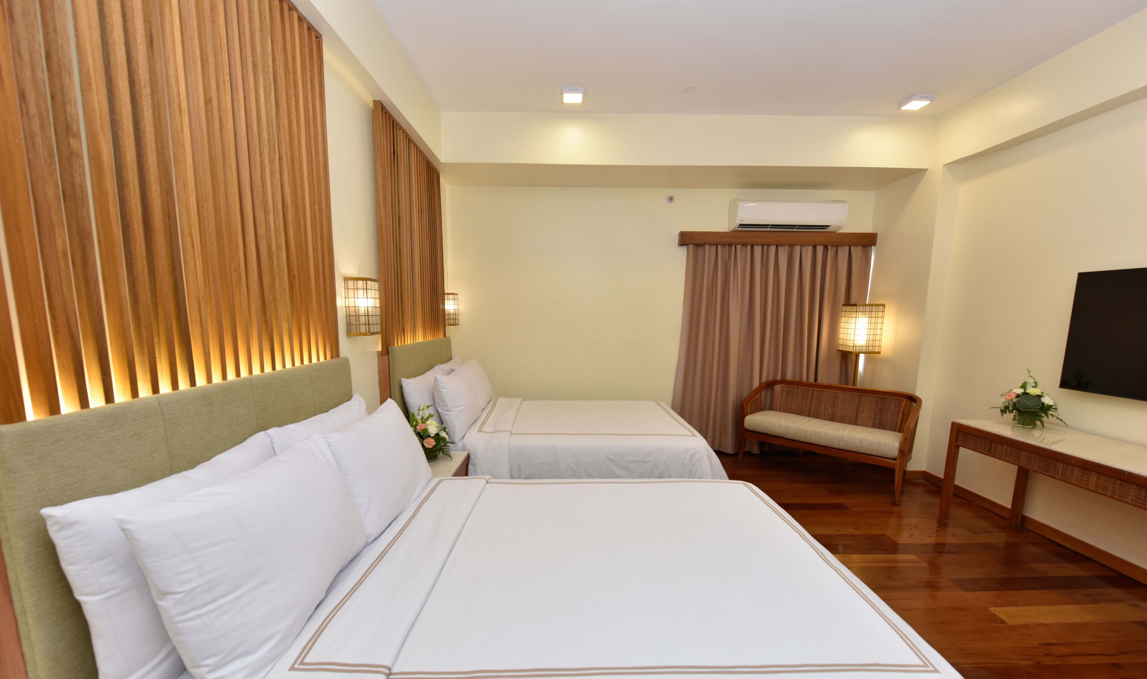 Habitación de hotel con dos camas, cabecero de madera, aire acondicionado montado en la pared, una mesita con lámpara y televisor en la pared.