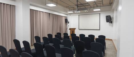 Un salón de conferencias con filas de sillas, un atril, una pantalla de proyector y cortinas.