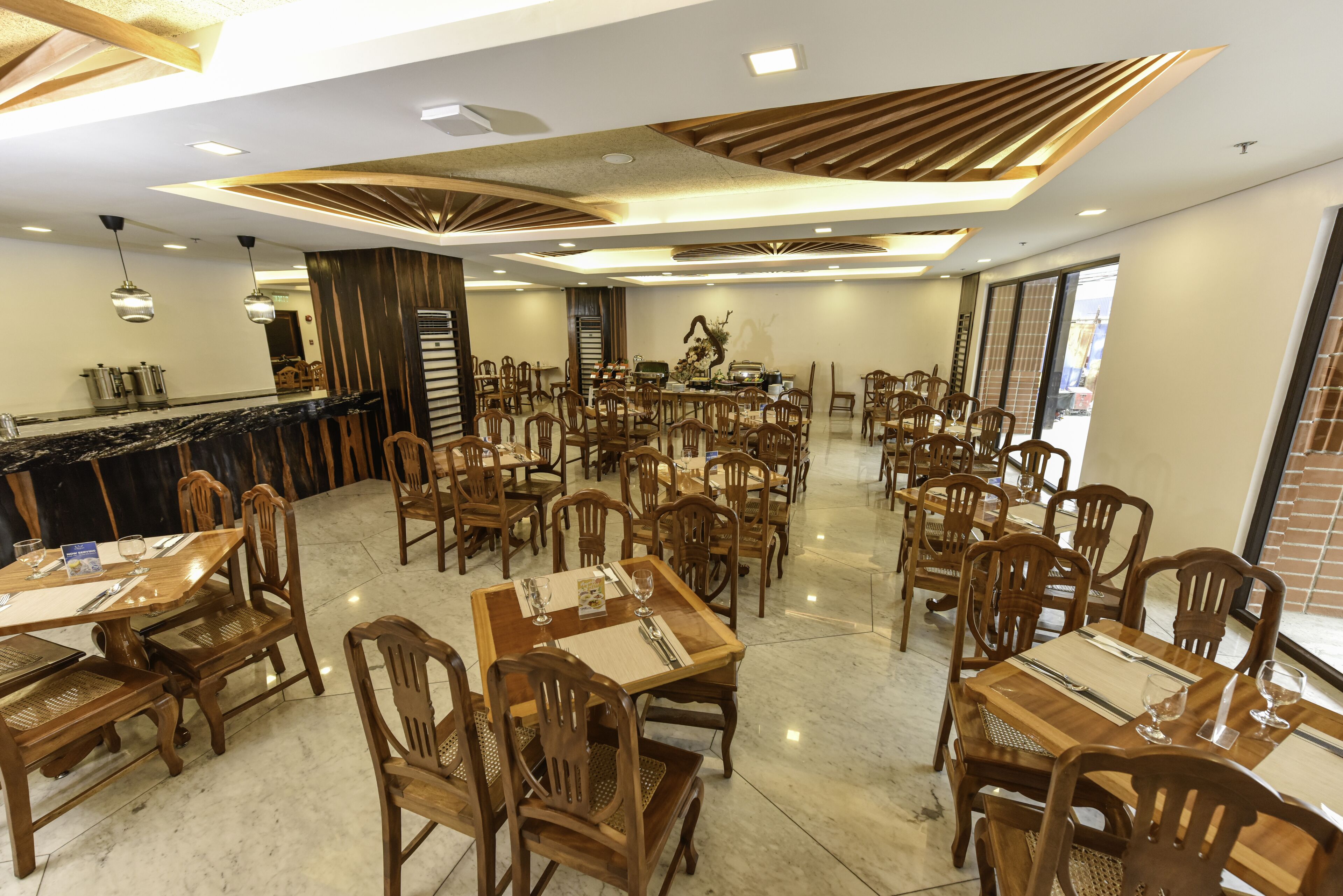 A spacious dining area with multiple wooden tables and chairs, a central bar area, and a modern ceiling design.