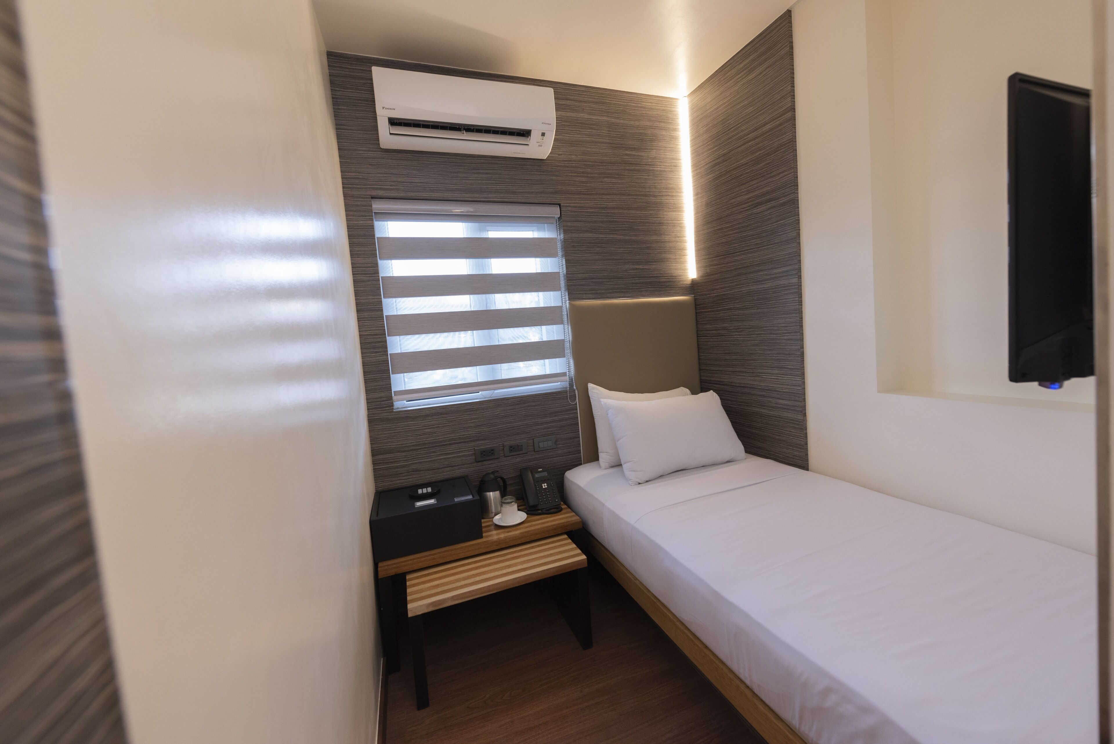 A compact hotel room with a single bed, a bedside table with a lamp, a small table with a phone and a coffee maker, and a window with blinds.