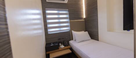 A compact hotel room with a single bed, a bedside table with a lamp, a small table with a phone and a coffee maker, and a window with blinds.