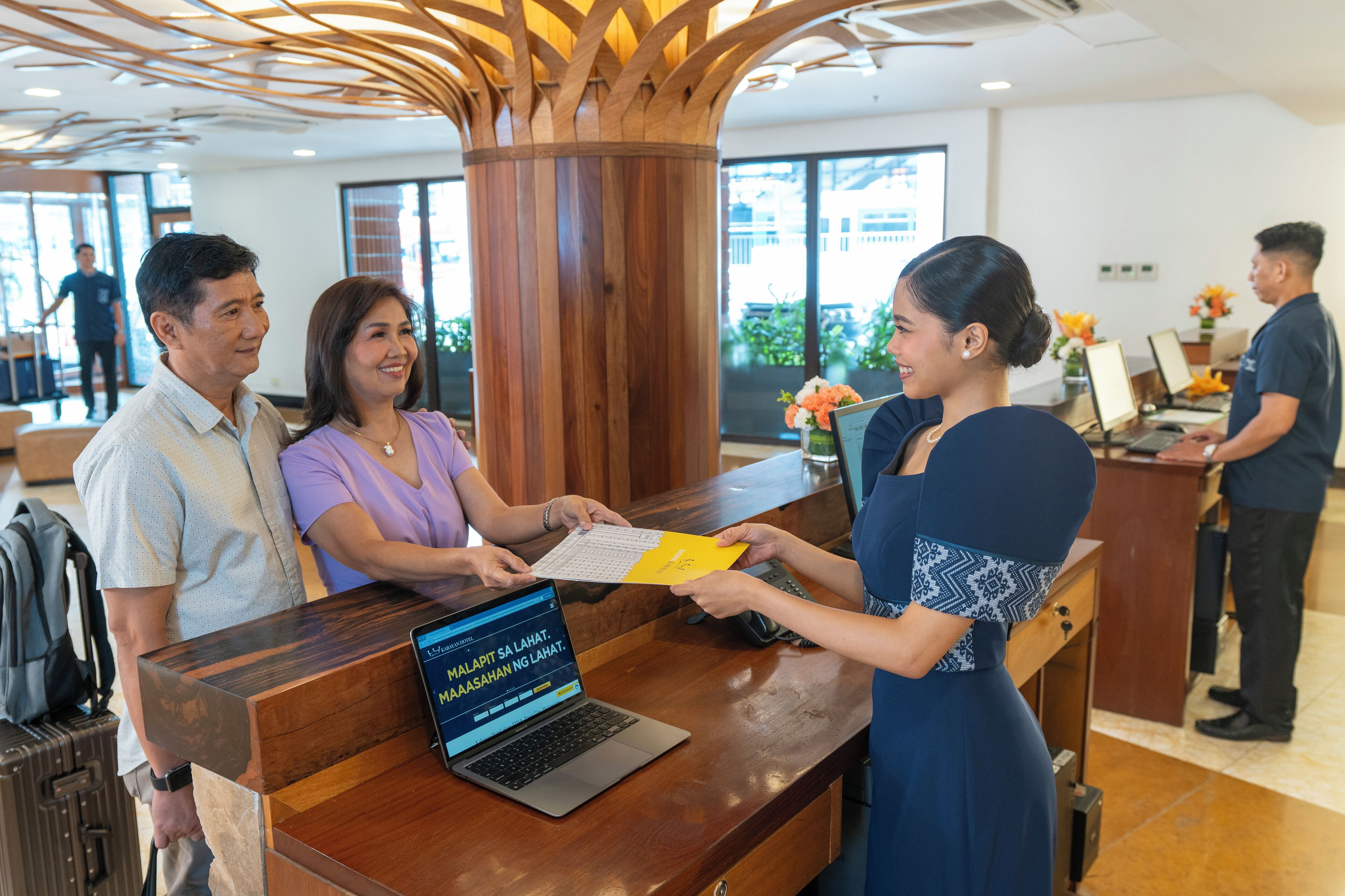 A reception desk with a laptop, a wooden pillar, and a person handing over a document.