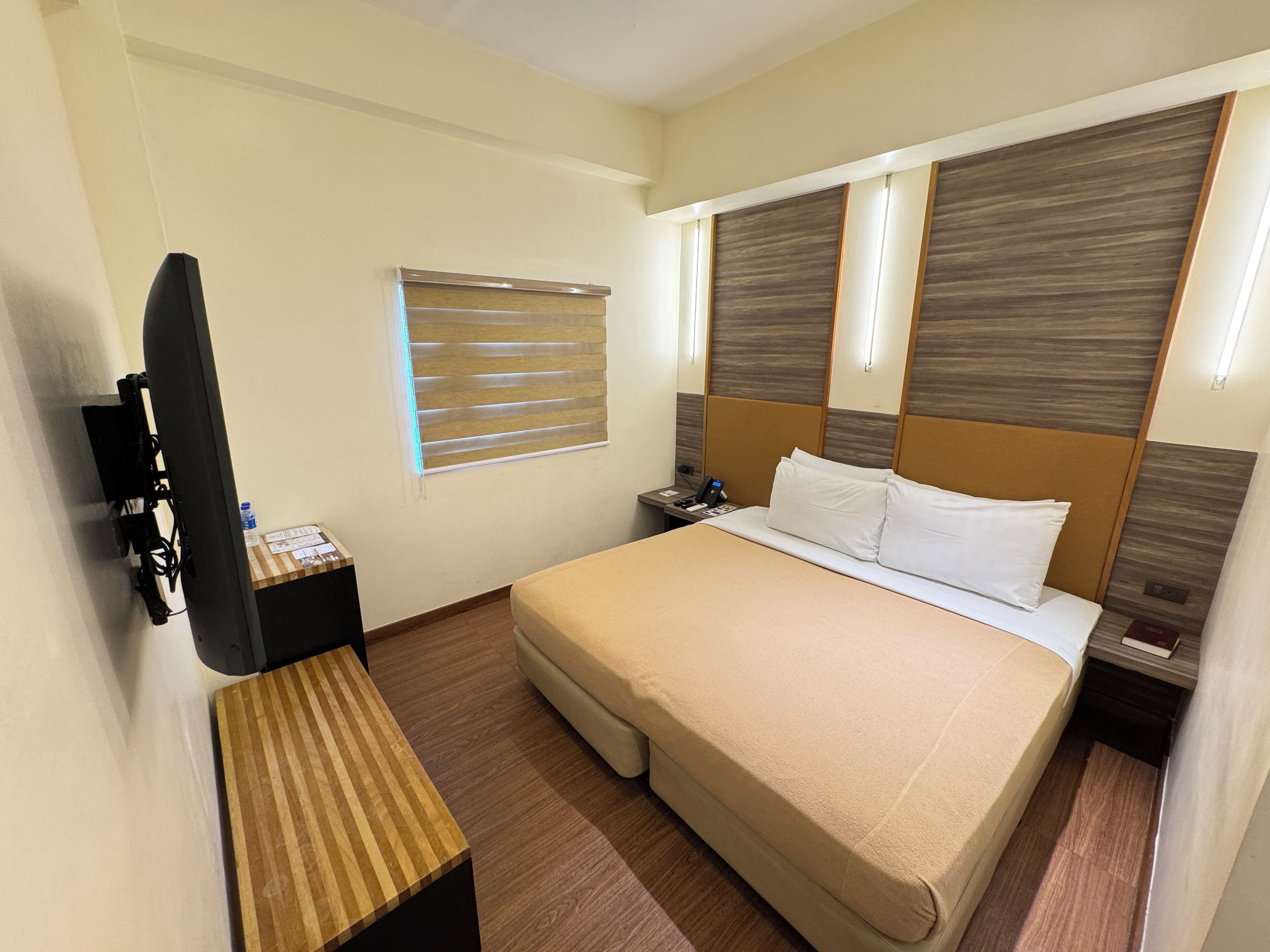 A hotel room with a bed, bedside table, TV, and a window with blinds.