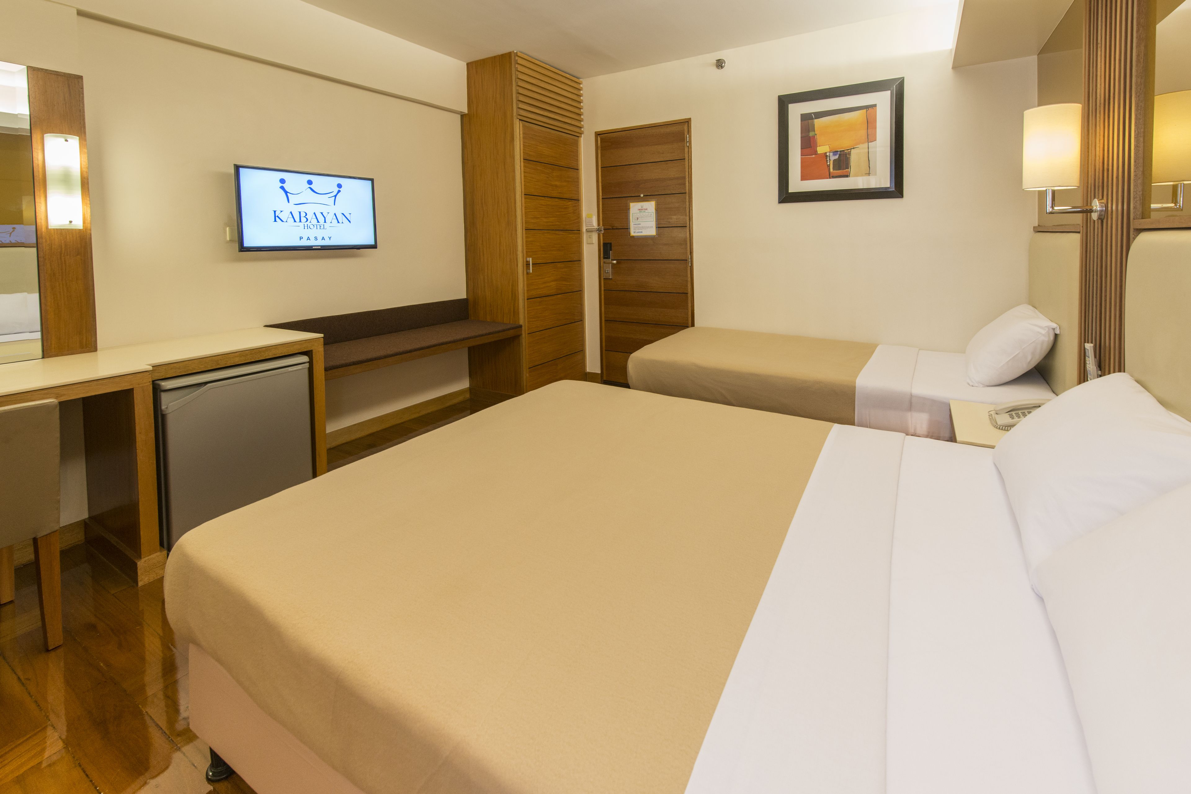 Habitación de hotel con una cama grande, un escritorio, un televisor mostrando el nombre del hotel y un armario de madera.
