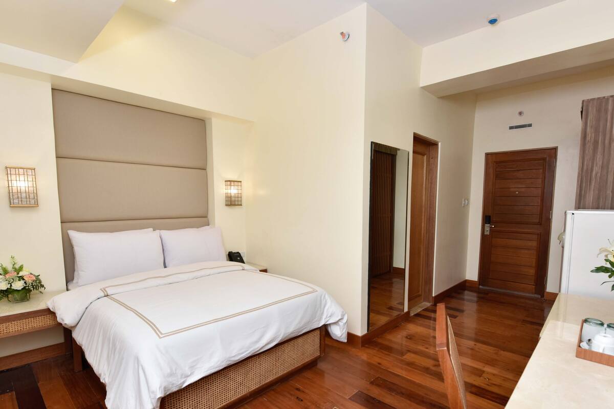A hotel room with a large bed, bedside table, and a small desk with a vase of flowers.