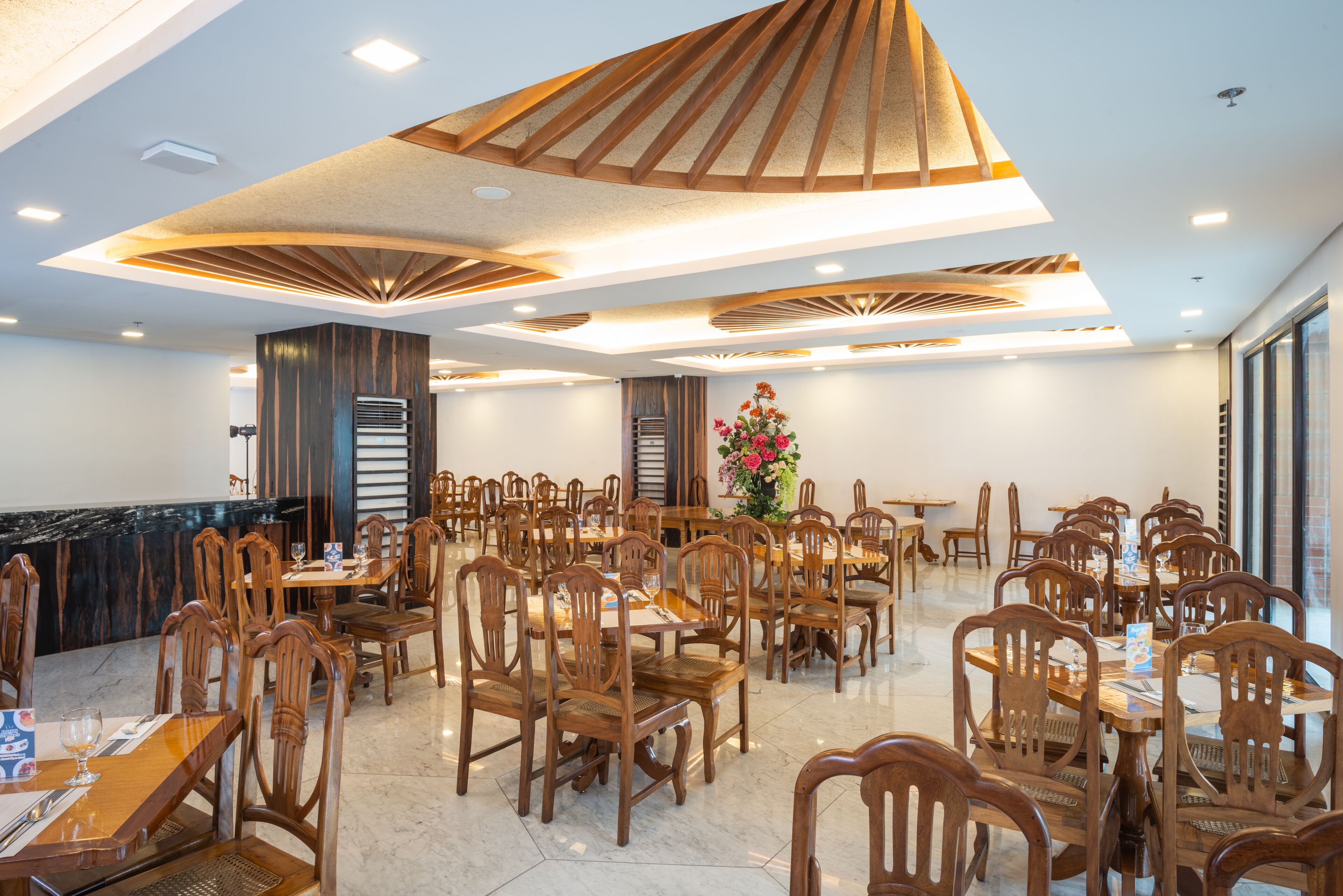 A spacious dining area with wooden tables and chairs, a central floral arrangement, and a modern ceiling design.