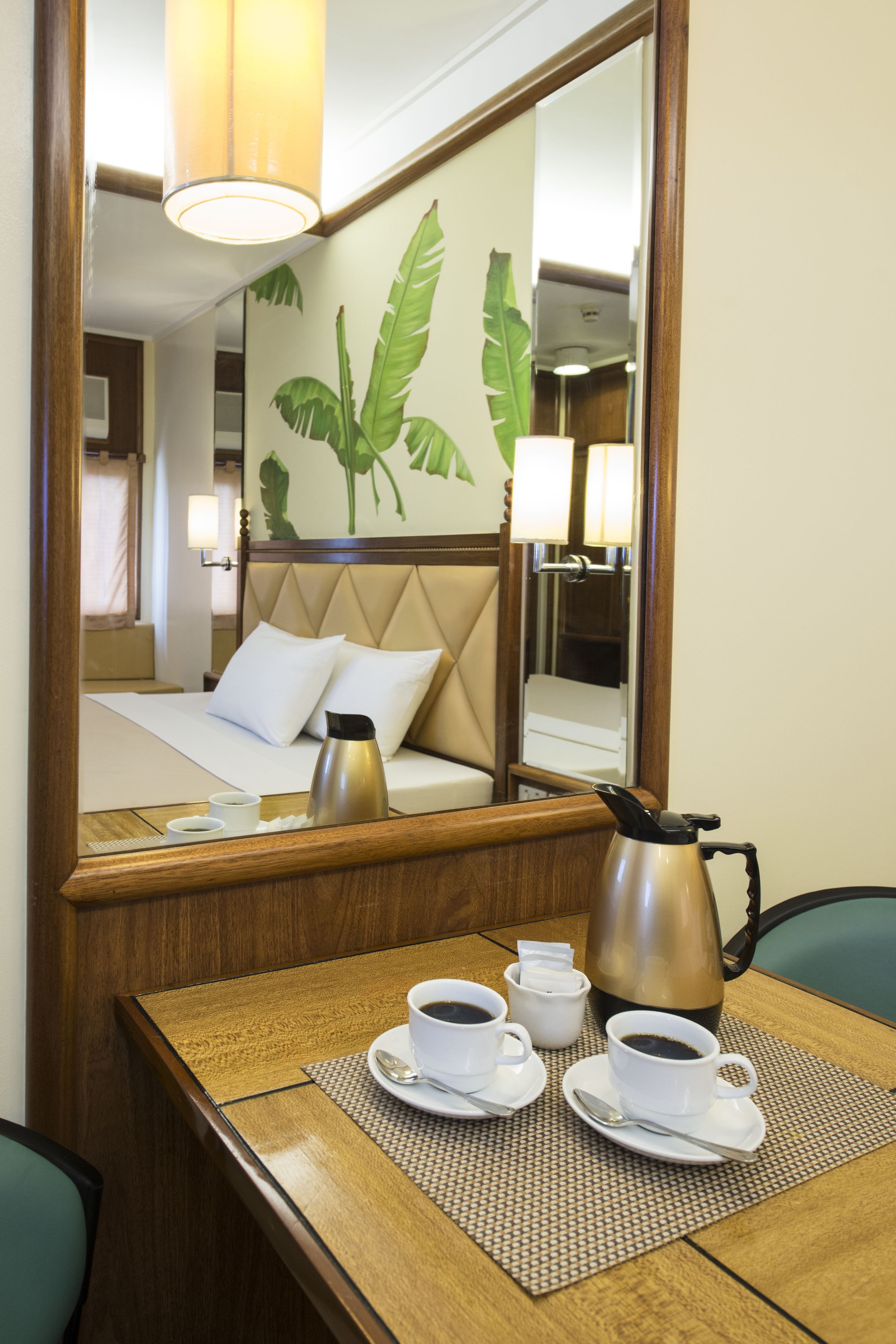 A hotel room with a bed, a wooden table with a coffee pot, cups, and a mirror reflecting the room.