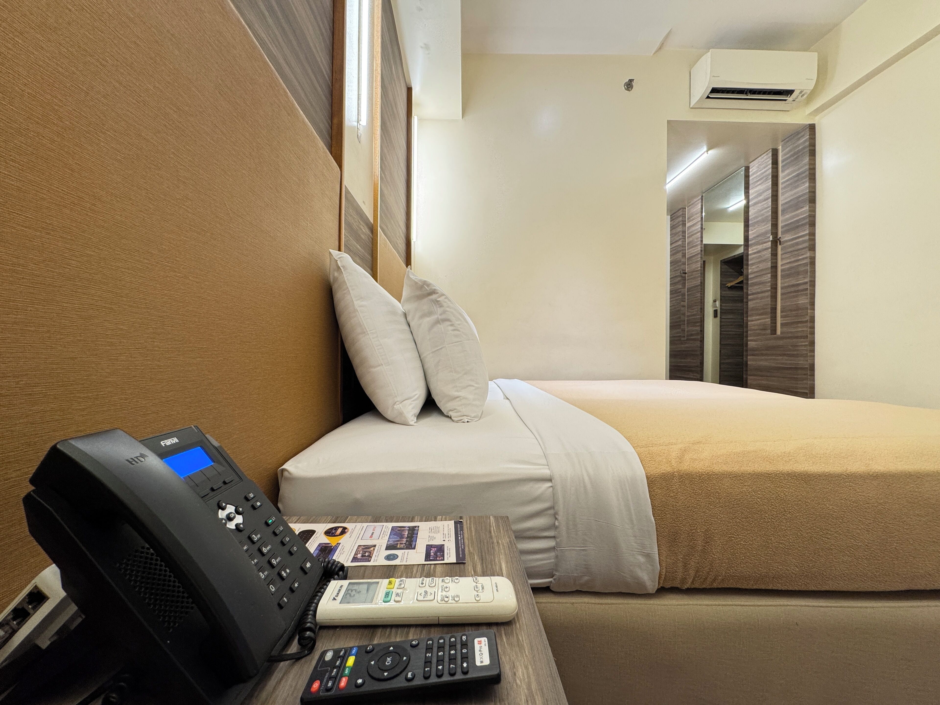 A hotel room with a bed, a bedside telephone, a remote control, and a wall-mounted air conditioner.