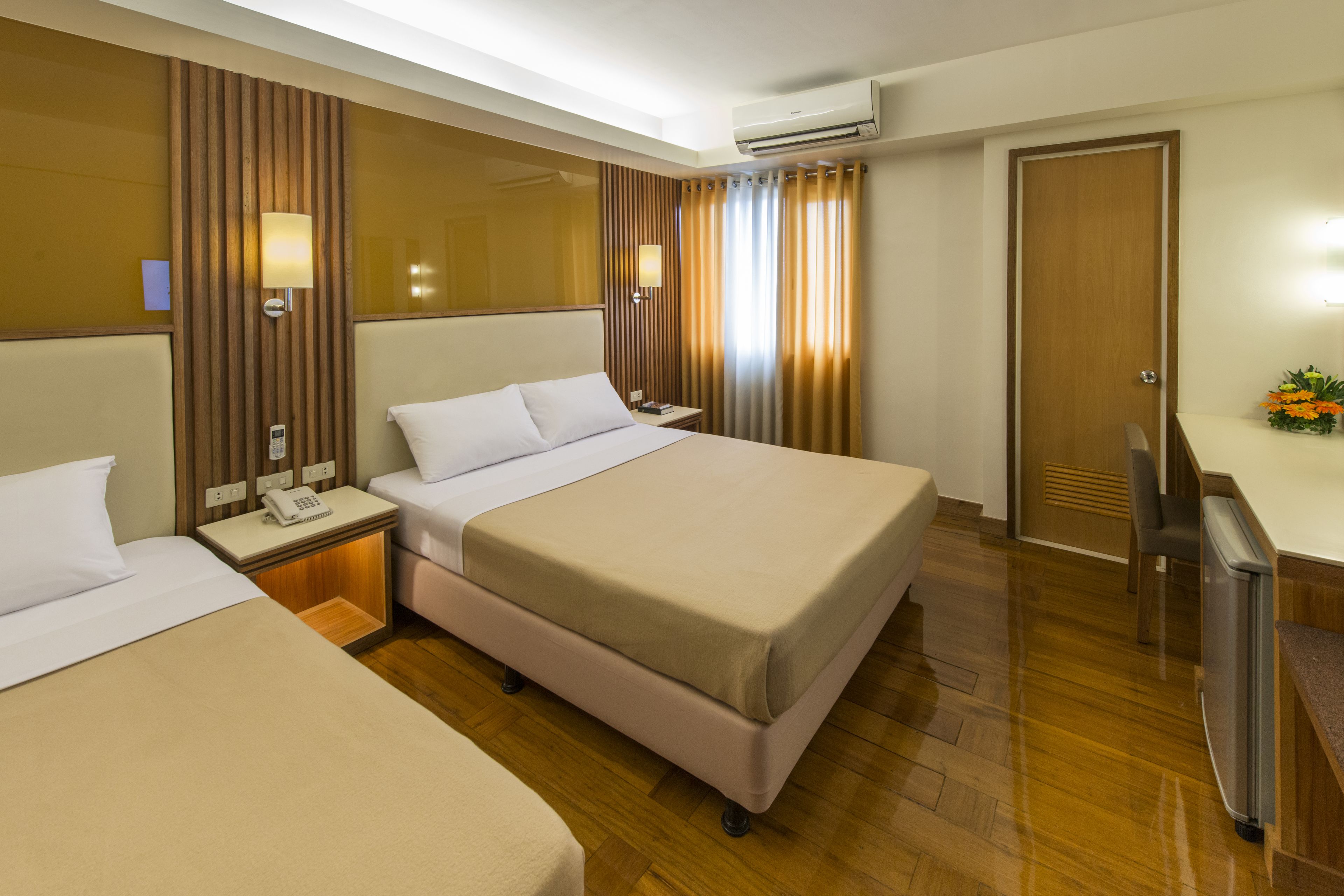 A hotel room with two beds, a desk, a chair, an air conditioner, and a small table with a vase of flowers.