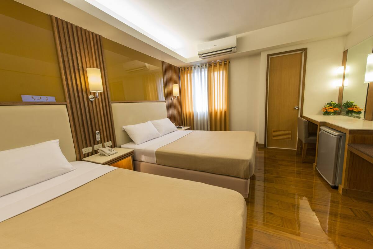 A hotel room with two beds, a desk with a telephone, a mirror, a small refrigerator, and a wooden floor.