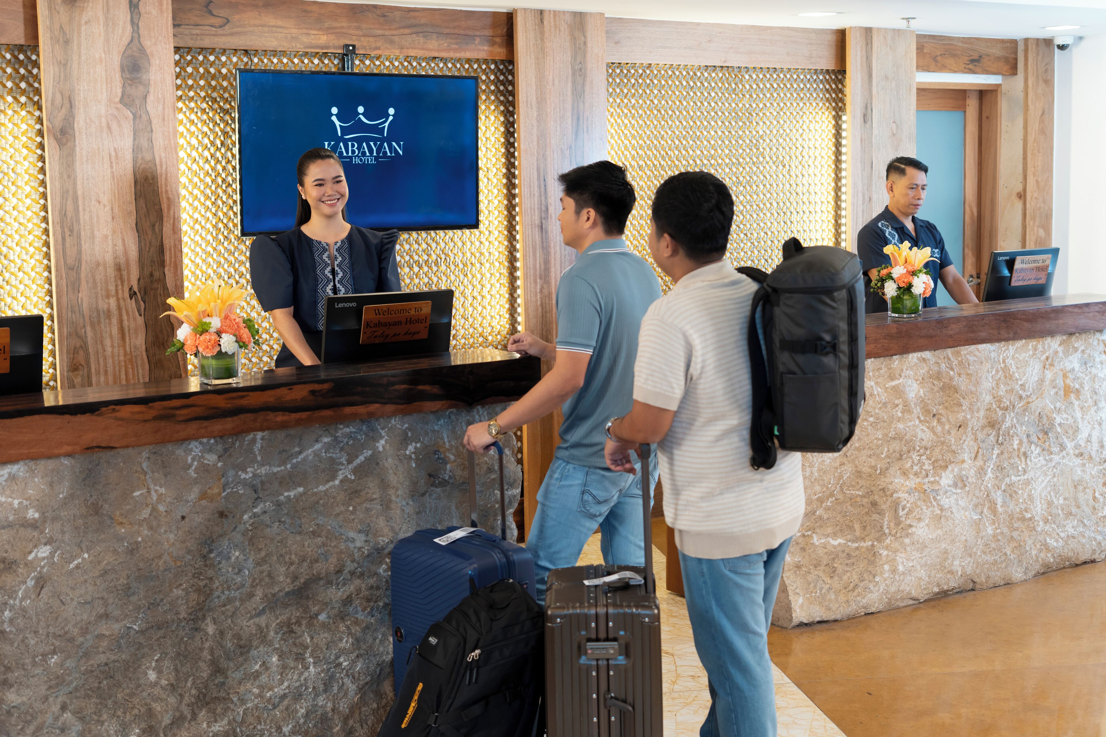 Mostrador de recepción del hotel KABAYAN con una pantalla de televisión mostrando el logotipo del hotel, un cartel de bienvenida y un jarrón de flores.