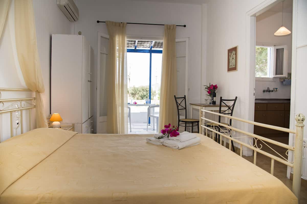 A hotel room with a bed, a desk, a chair, a small table with a vase of flowers, and a view of the outside.