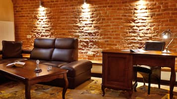 A cozy living room with a leather sofa, a wooden coffee table, and a desk with a lamp. A brick wall with a light fixture is visible in the background.