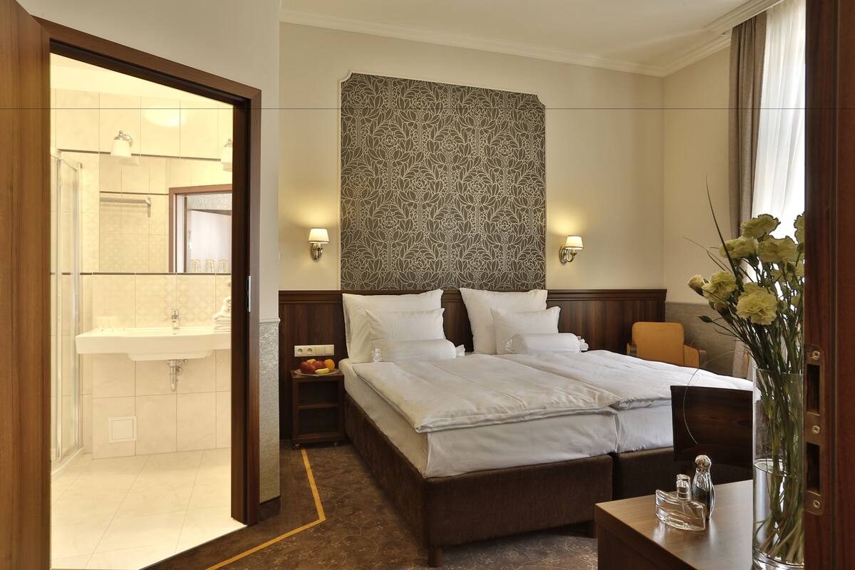 A hotel room with a large bed, a nightstand with a lamp, a vase with flowers, and a view of the bathroom.