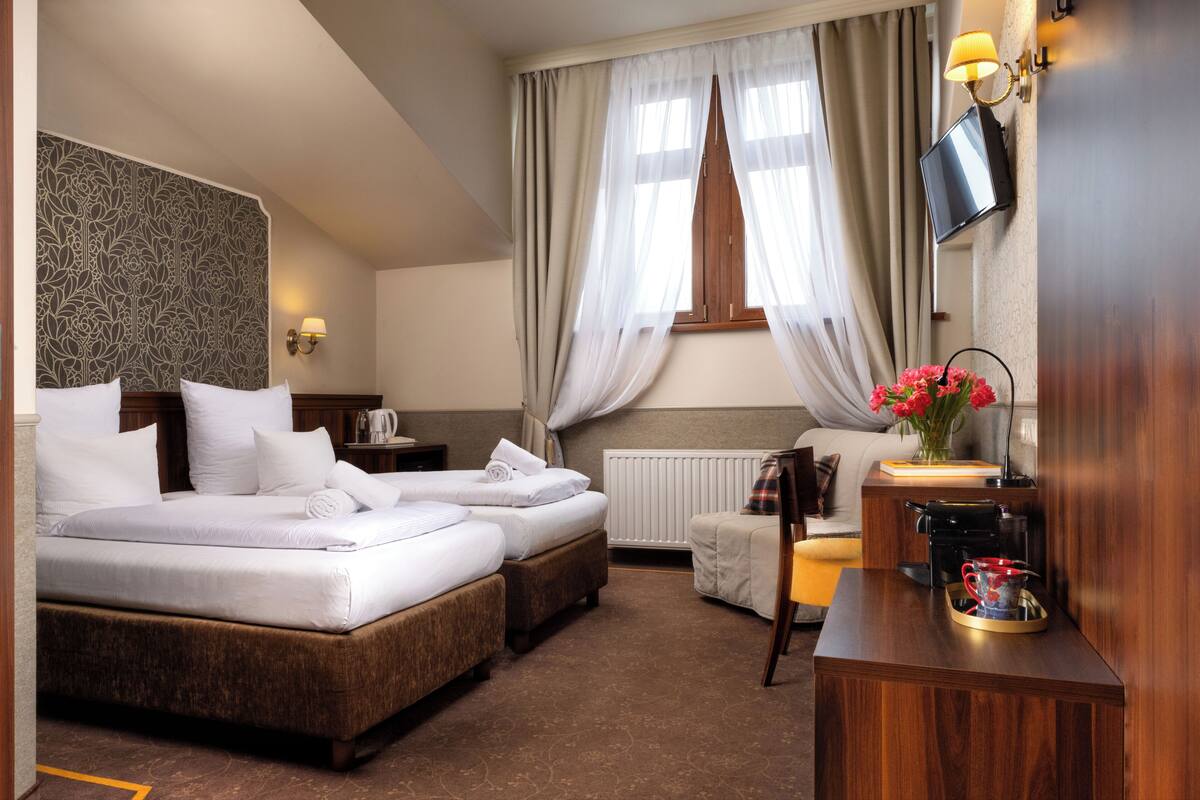 A hotel room with two beds, a sofa, a small table with a lamp, a TV, and a window with curtains.