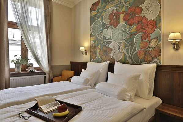 Une chambre d’hôtel avec deux lits, un grand décor mural représentant des motifs floraux, une fenêtre avec des rideaux légers, une table de chevet avec une lampe et un plateau contenant un livre, des verres et des fruits.