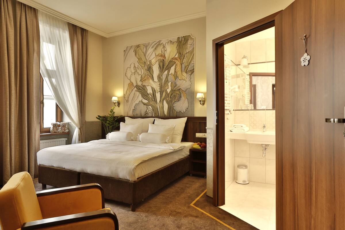 A hotel room with a large bed, a bedside table, a nightstand, a chair, a wall-mounted lamp, a painting, and a bathroom visible through an open door.