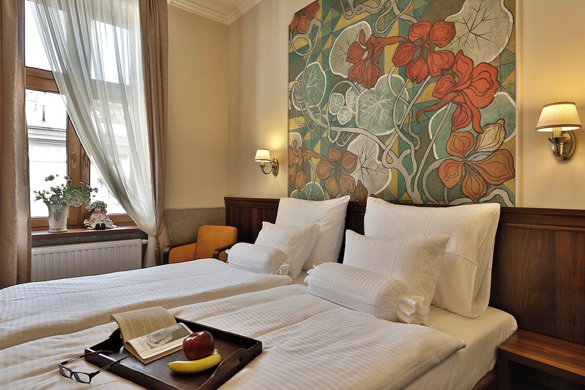 A hotel room with two beds, a window with curtains, a floral wall mural, a nightstand with a lamp, and a tray with a book and fruit.