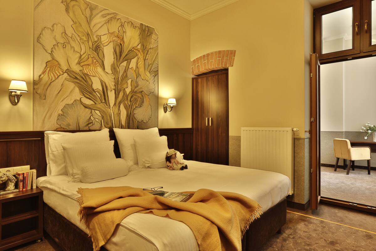 A hotel room with a large bed, a nightstand with books, a wall mural, and a view of the dining area through an open door.